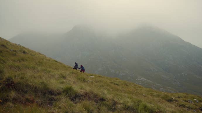 Un par de concursantes de 007: Road to a Million suben una colina empinada en Escocia, bajo la sombra de una montaña más grande y cubierta de nubes.