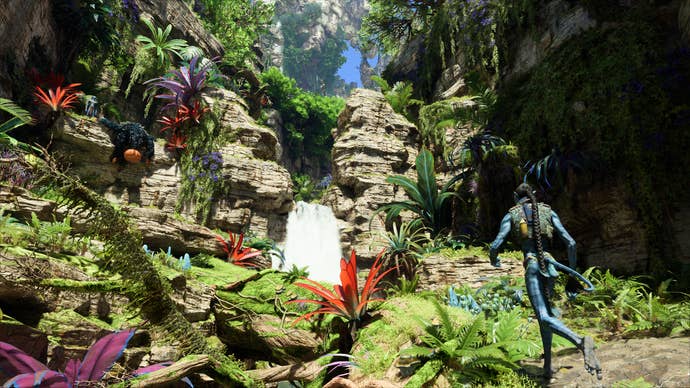 Avatar: Fronteras de Pandora: una cascada en la jungla, colores vibrantes, verdes intensos y rojos intensos, mientras un Na'vi admira el paisaje.