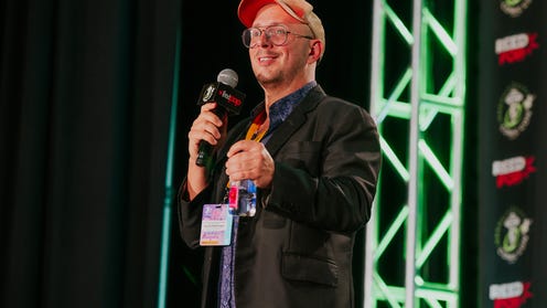 Steve Burns wearing a cap, glasses, and holding a microphone on stage