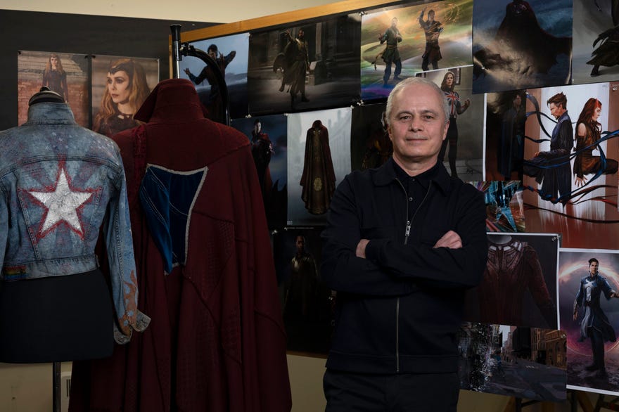 Costume designer Graham Churchyard (a white male wearing all black, 838-style) poses in front of the costume design board for Doctor Strange in the Multiverse of Madness. Also pictured is Strange's maroon Cloak of Levitation with a navy blue patch and America Chavez's Balenciaga jacket with a rainbow flag and star on the back.