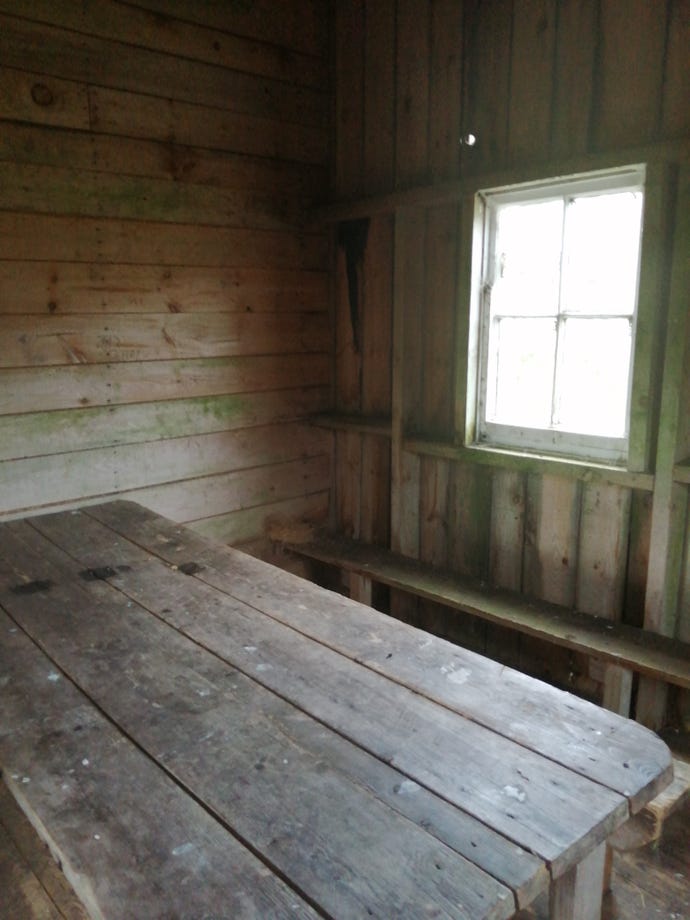 The inside of a hut for hikers in Scotland, showing a bare wooden table and a window with bright daylight outside