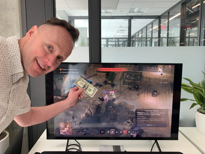 Harebrained Schemes' Chris Rogers stands in front of a TV holding a signed dollar, which he won in a bet during our GDC Lamplighters League preview session