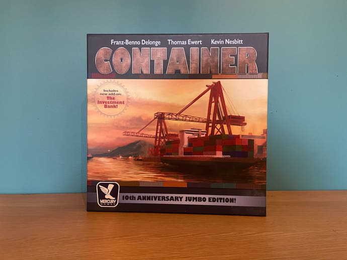La caja del juego de mesa Container (Jumbo Edition), que es un juego sobre barcos portacontenedores.  Muestra una imagen en tonos de puesta de sol de un barco portacontenedores cargando.  Brillante.