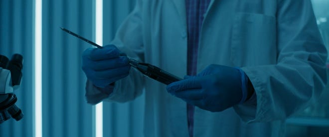TV still of scientist character wearing a lab coat and gloves and holding a very large needle