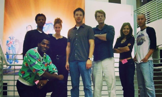 A group of voice actors standing together with a large Justice League poster in the background