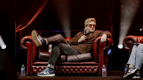 Photograph of Rhys Darby reclining on a couch onstage at MCM