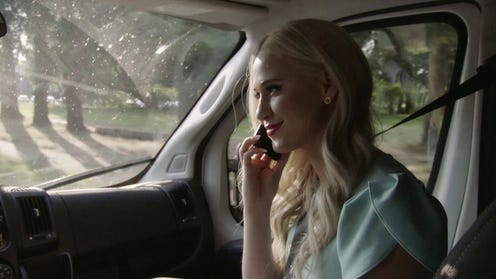 A person with blonde hair riding in the passenger seat of a car and listening to a phone. Maria Bakalova in Borat Subsequent Moviefilm