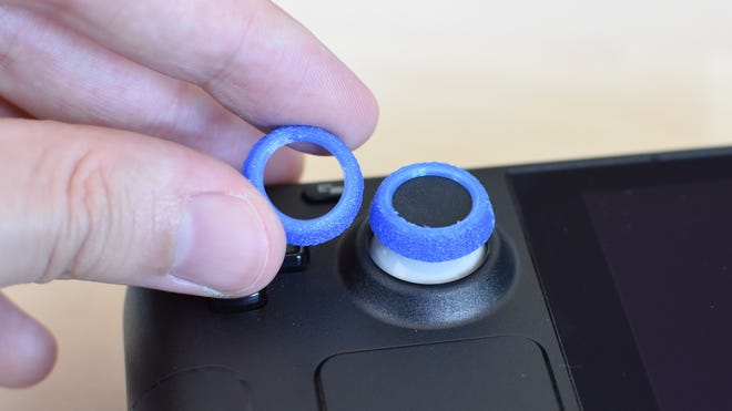 The NinjaPrint3D Steam Deck Thumb Grips. One is applied to the Steam Deck's left thumbstick, the other is being held next to it.