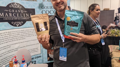Joe Peck holding a bag of coffee and a bag of cocoa at the Grand Marceline booth