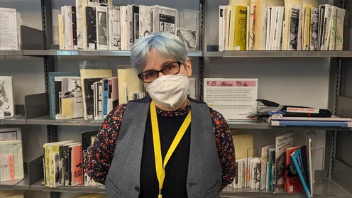 Zine librarian Jenna Freedman with short colored hair, wearing glasses, a mask, and a yellow lanyard in front of shelves featuring zines