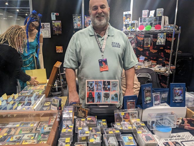 Photograph of a man wearing a polo shirt standing  in a booth selling Star Wars collectable cards