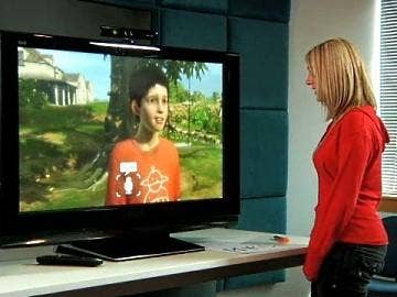 Screen of the Project Milo E3 Kinect demo video with a young boy in an orange shirt on the TV talking in the direction of a woman in a red sweater standing in front of the TV.