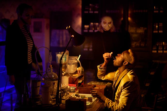 A picture showing a manly man in a suit taking a drop of some unknown liquid while masked men, in a dark room around them, watch.