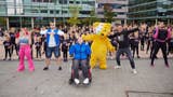 A crowd of people dance outside in a city square, including Pudsey the Bear, the Children in Need mascot.