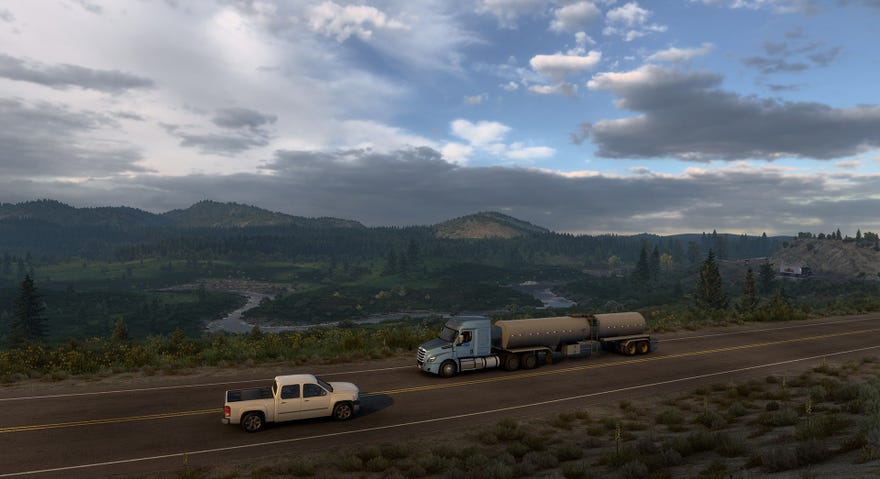 A screenshot of American Truck Simulator's Wyoming DLC showing a truck driving on a road in front of a sprawling vista of river, mountains and trees.
