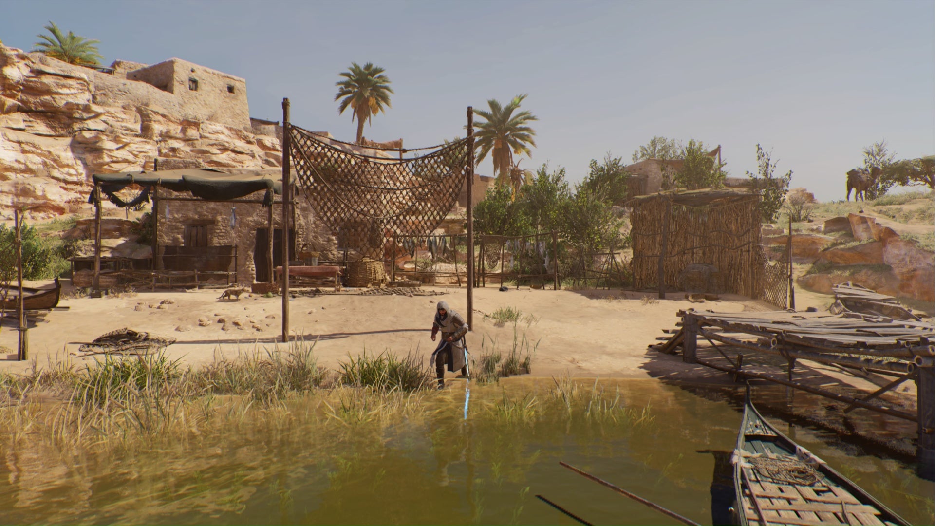 Assassin's Creed Mirage, una toma amplia de Basim parado en la orilla de un río con un bote a la derecha, redes de pesca y peces colgando de postes de madera detrás de él, además de una casa y una cabaña a ambos lados de Basim en la distancia.