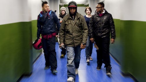 A still image of five young men in street clothing walking down a lit hallway