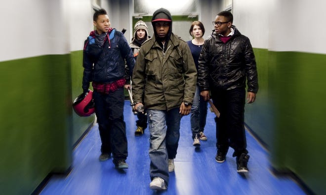 A still image of five young men in street clothing walking down a lit hallway