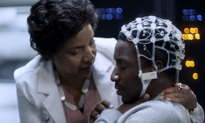 A still image of a woman in a white suit holding onto a man wearing a grey sweatshirt and a medical equipment hood