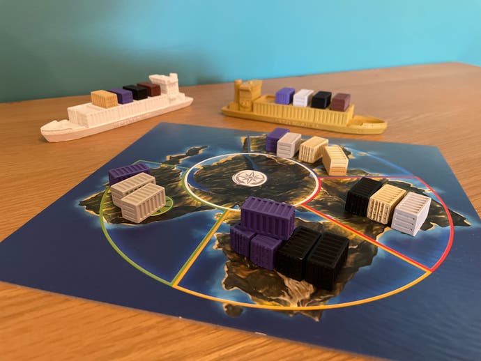 Another board from the Container board game (Jumbo Edition), showing a geographical region - possibly the world - and se around it. It's divided up into segments and there are palm-sized shipping container pieces placed in each. Two container ship playing pieces wait on the table outside of the board's area.