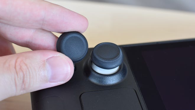 The Dbrand Steam Deck Stick Grips. One is applied to the Steam Deck's left thumbstick, the other is being held next to it.