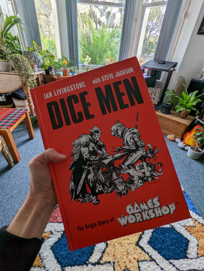 Bertie's hand, and adjoining arm, holding up the Dice Men Games Workshop history book. It's bright red with two characters - one from fantasy, one from sci-fi - playing a game of dice together. Bertie's flat can be pictured in the background. It has some plants in it.
