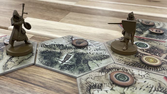A pair of small figurines are placed on top of hexagonal game-world tiles on a wooden table. There are a couple of tokens on top of them too. It looks like someone is having a jolly good board game time.