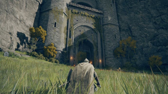 An armoured character wearing a cloak in grass crouching outside of a castle's entrance in the Limgrave area in Elden Ring.