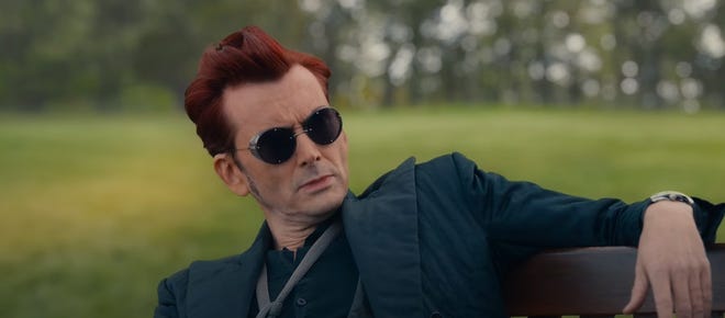 Still image of David Tennant as Crowley sitting outside on a bench