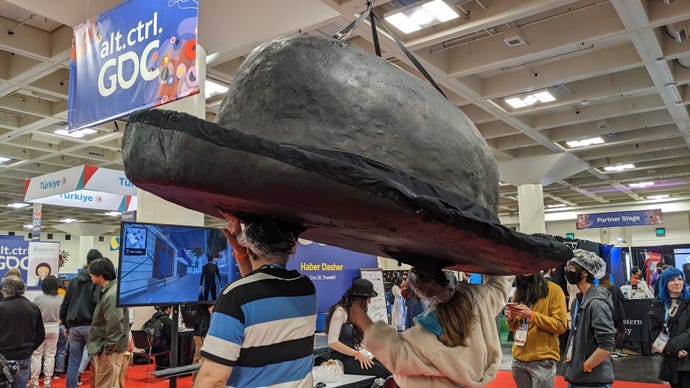 Two people stand underneath a giant black bowler hat suspended from the ceiling at GDC