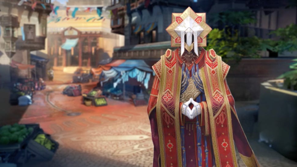 A citizen of Meridian, one of the largest cities in the world of Horizon Zero Dawn, stands in the foreground with their hands clasped. They wear red robes and five-pointed mask shaped like a sunburst.