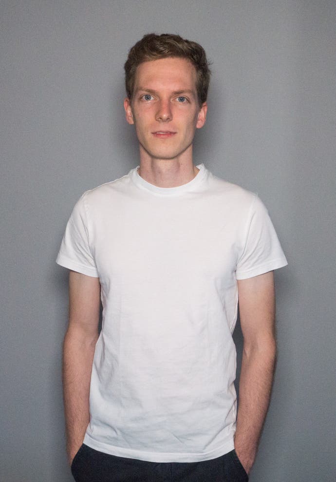 A portrait of Philip Stollenmayer, with short hair and an enigmatic smile. He is wearing a white T-shirt and has his hands in his pockets.