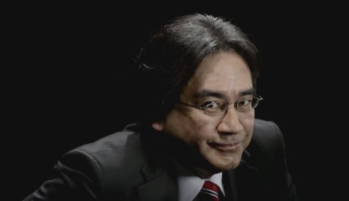 Close-up picture of Satoru Iwata in a suit in a dark room. He is in a half-crouch position and has a mischievous smile on his face