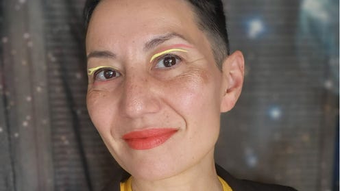 Profile photo of MariNaomi, a queer Japanese American person with light skin and shorn black hair, wearing a dark blazer over a yellow t-shirt that says "Asian Love" in red-orange. They are posed in front of a dark and starry outer space backdrop.
