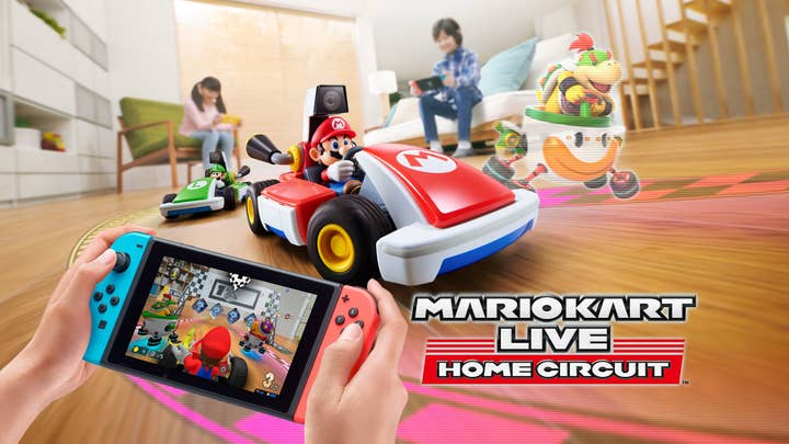 A promo pic for Mario Kart Live Home Circuit showing a Mario RC car speeding through a living room with AR-added track elements around it. In the foreground, hands hold a Switch showing a camera view from atop the RC car