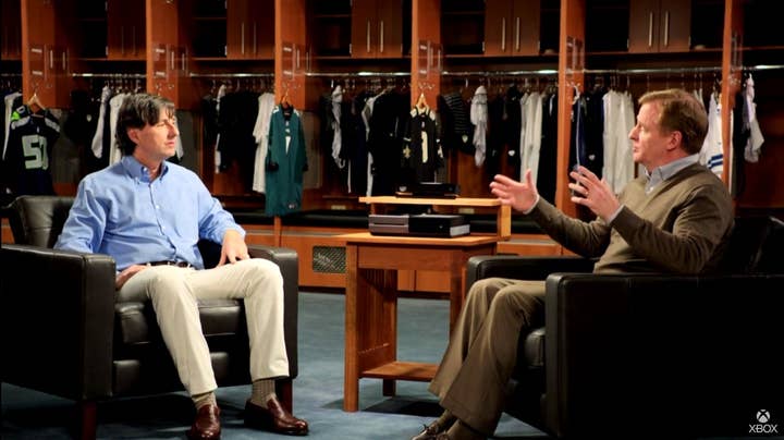 Don Mattrick and Roger Goodell talking while sitting in large black chairs in a locker room. An Xbox One sits on a small table placed between them