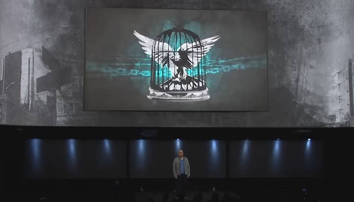 A man on stage with a giant screen behind him showing a picture of a bird with a cage transposed over it and chains and barbed wire behind it