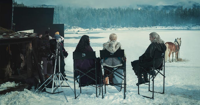 A production shot of season three of The Witcher, showing the actors and actresses behind Geralt, Ciri and Yennefer chilling in the snow on those tall actor chairs.