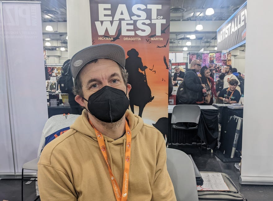 A photograph of Nick Dragotta wearing a mask and a tan hoodie in front of an East of West banner at New York Comic Con