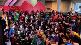 A large group of people, some wearing costumes of their favourite video game characters, gather around a large PAX logo to take a photograph together.