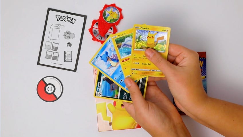Uma mão divulga quatro cartões Pokemon TCG encontrados na mais nova promoção do McDonald's Meal. Na mesa, há um cartão de instrução, uma moeda e um girador de plástico