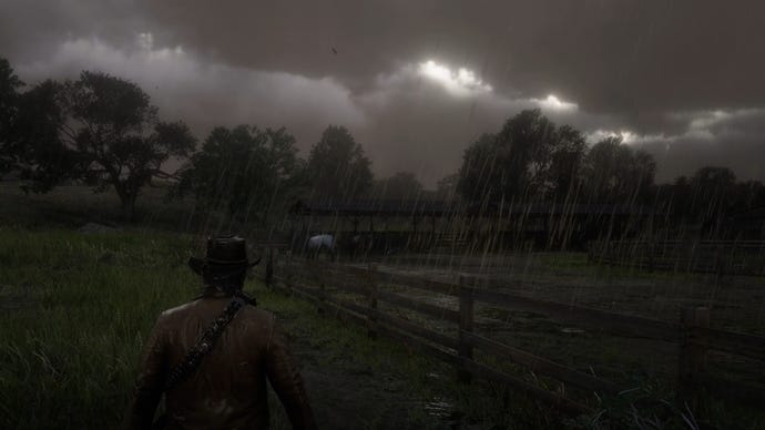 A screenshot from Red Dead Redemption 2 of Arthur walking in the rain next to a field.