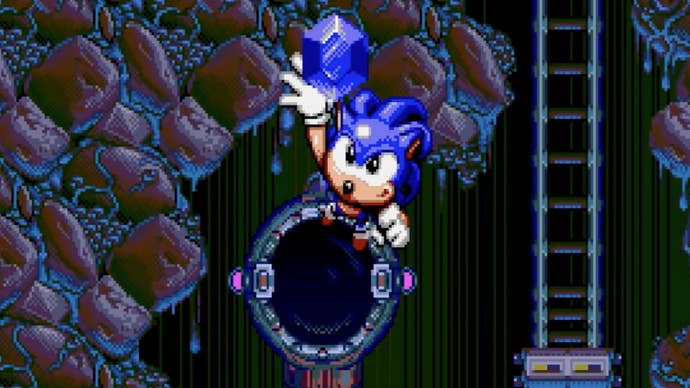 Sonic, in his Spinball incarnation, reaches towards the screen after jumping out a hole, holding a Chaos emerald.