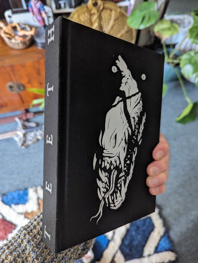 A photo of Bertie's hand holding a thick black hardback book with the a monster's shadow-covered face on the front, and the words "Teeth" on the spine.