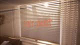 A screenshot from the interior of an apartment, angled towards the pulled blinds on a large window which are letting sunlight through the slats. The more eye-catching thing, however, is the "Don't Trust" slogan spray painted on top of them.