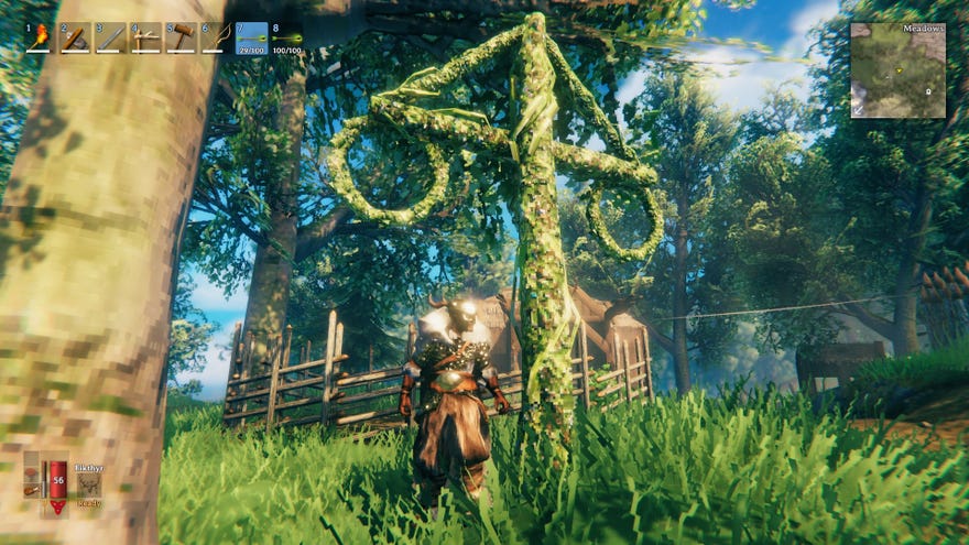 Valheim - A player in armor standing next to a maypole covered in greenery inside a small village.