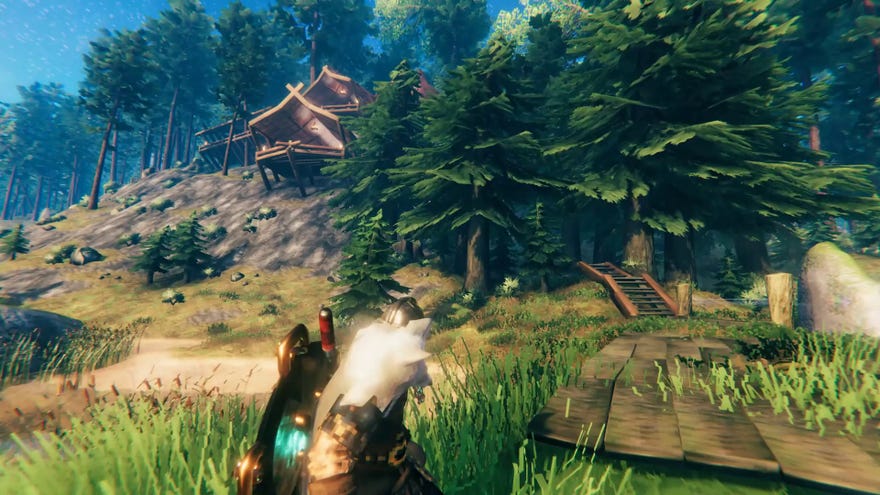 A Valheim screenshot which shows a player looking out at their home perched on the rocks above. A wooden path leads through pine trees towards the entrance.